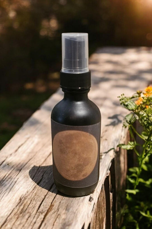 A matte black spray bottle with a clear cap and a blank circular label, resting on a weathered wooden fence outdoors with green foliage and yellow flowers in the background.
