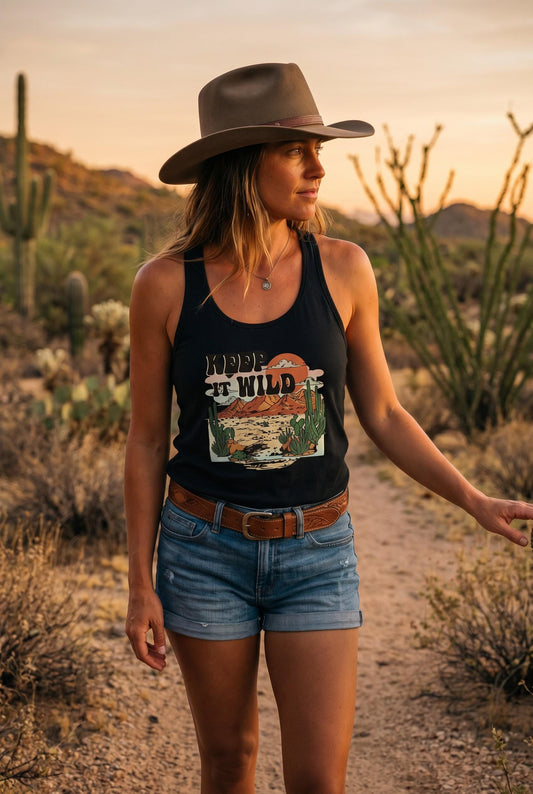 A woman wearing a black racerback tank top with a western-themed desert graphic that reads 'RIDE IT WILD', paired with denim shorts and a brown hat, standing on a dirt path in a desert landscape at sunset.