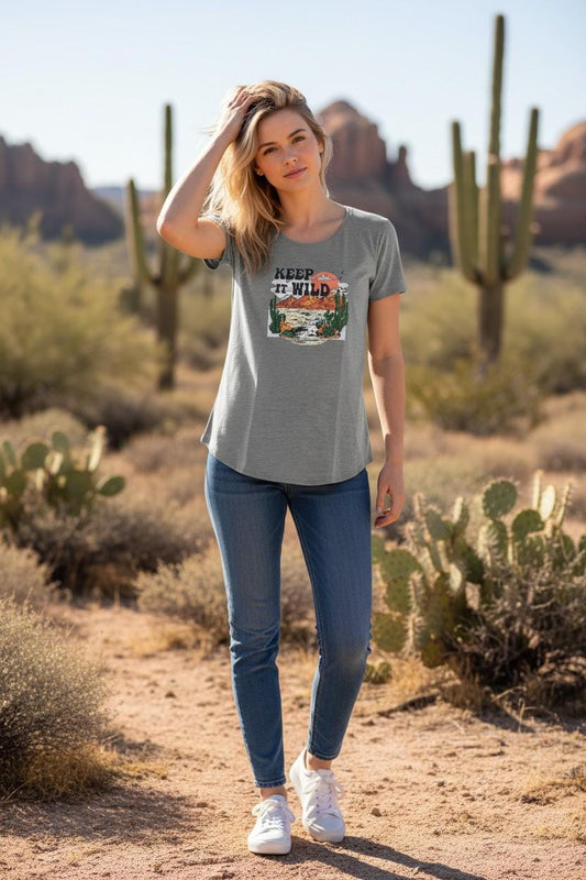 A woman stands on a desert trail wearing a heather gray graphic t-shirt with a 'KANSAS CITY WILD' sports print, blue jeans, and white sneakers, with cacti and rock formations in the background.