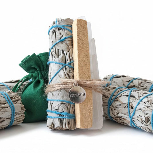 A set of dried white sage smudge bundles wrapped in blue string, with one bundle tied in front with twine and a round silver tag reading 'dream'. A small green fabric drawstring pouch sits behind the bundles on a clean white background.