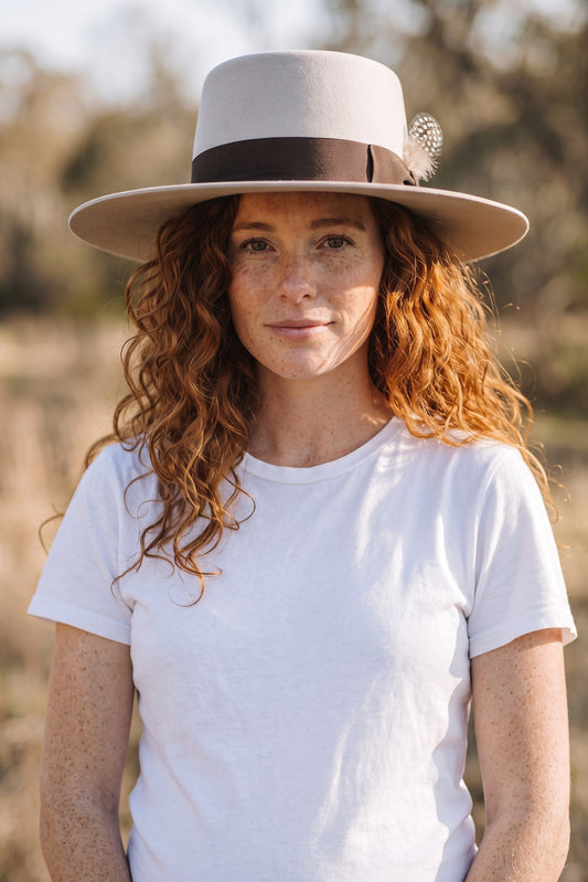 Spirit Warrior Provisions sand gambler hat with flat brim and grosgrain ribbon worn by a woman with curly red hair.